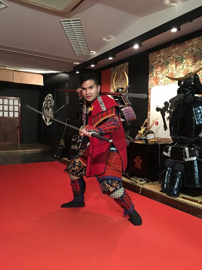 Nico dressed in red samurai armor, holding a katana, posing in a studio with more armor on display behind him.