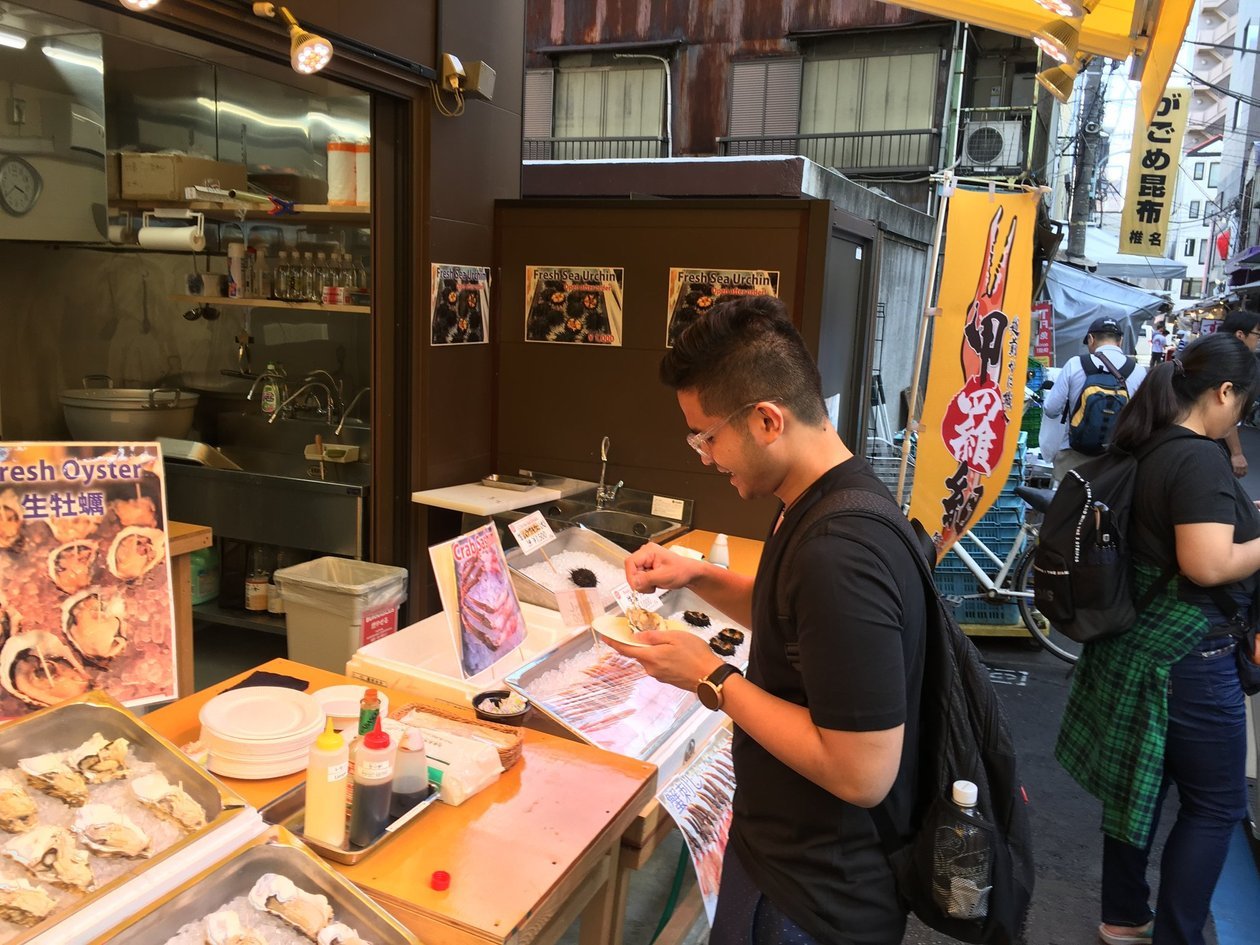 Nico in a black t-shirt eating sea urchin from a paper plate at a fish-market stall, with oysters on ice beside him.