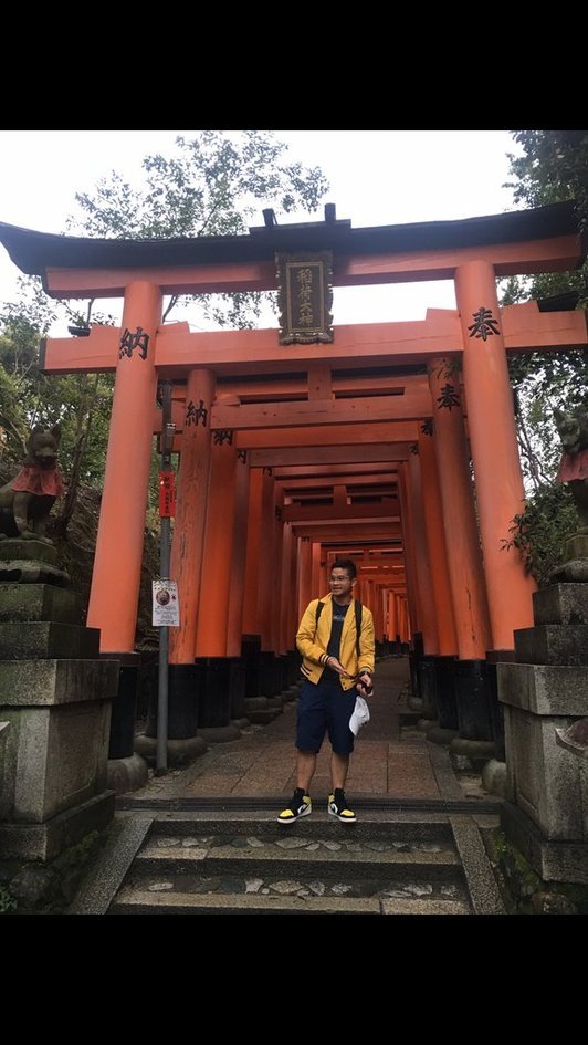 Nico in a yellow jacket and blue shorts standing on the steps under the orange torii gates at Fushimi Inari.