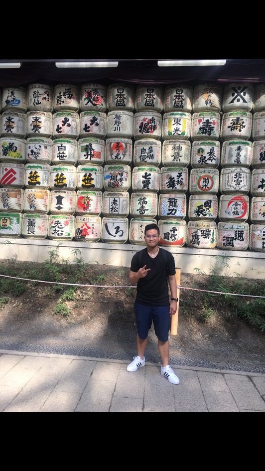 Nico in a black t-shirt standing in front of a wall of stacked sake barrels covered in Japanese calligraphy.