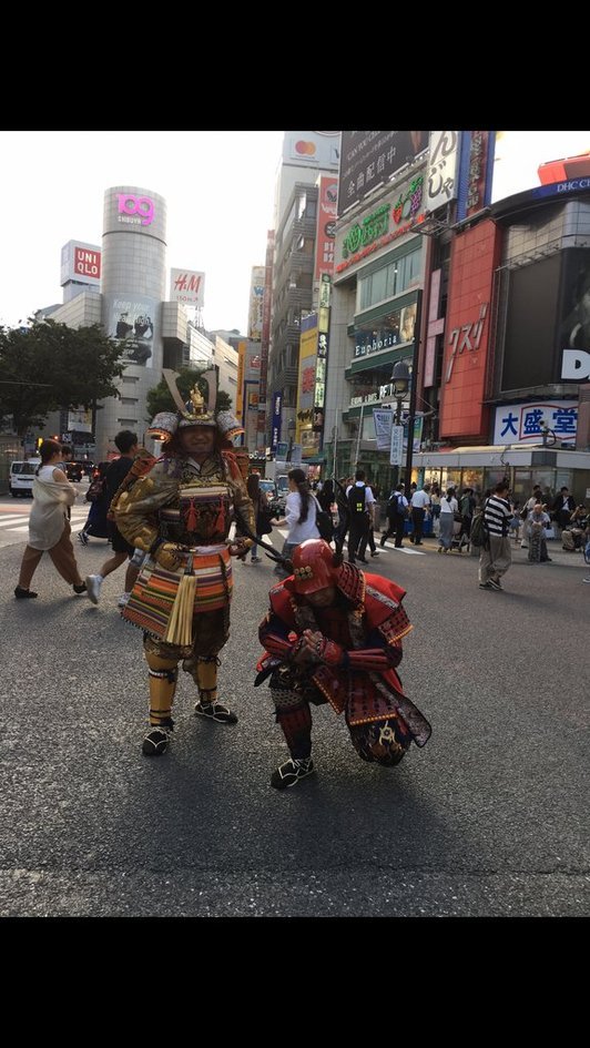Two street performers in samurai armor posing in a Shibuya intersection, with the 109, Uniqlo, and H&M buildings behind them.