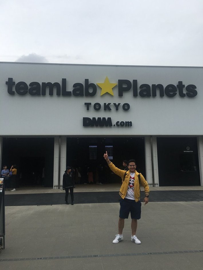 Nico in a yellow jacket pointing up at the teamLab Planets Tokyo sign on the white museum facade.