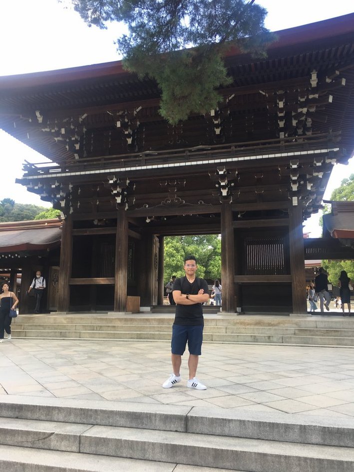 Nico standing with arms crossed in front of a dark wooden Japanese temple hall, a pine branch hanging over the roof.