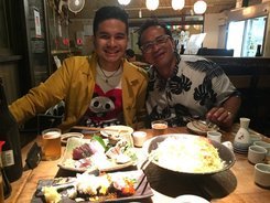 Nico in a yellow jacket and Dad in a leaf-print shirt smiling over a hot pot, sashimi, and sake cups.