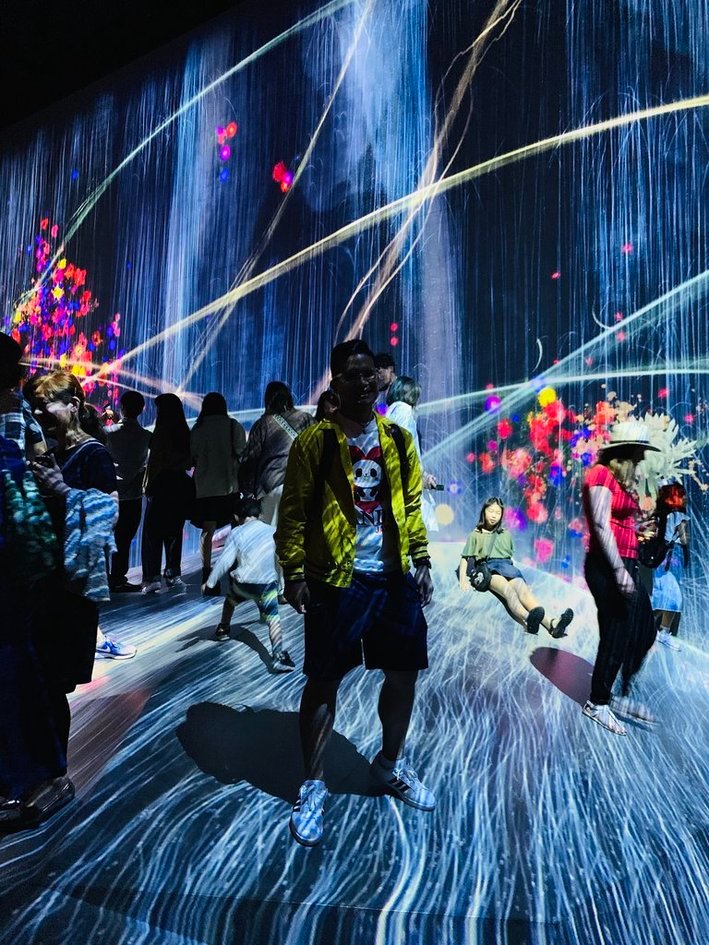 Nico in a yellow jacket standing in a dark teamLab room where blue light and flowers are projected across the floor and walls.