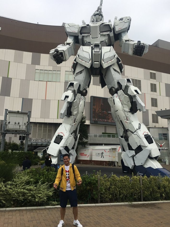 Nico in a yellow jacket standing in front of the life-size Unicorn Gundam statue outside DiverCity Tokyo Plaza in Odaiba.