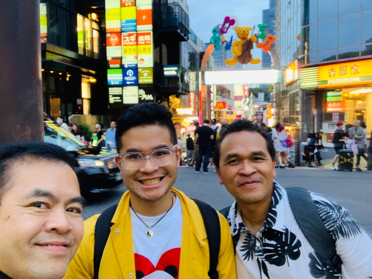 Selfie of Dad’s friend, Nico in a yellow jacket, and Dad in a leaf-print shirt on a bright Tokyo street, with a Yoshinoya and a bear-shaped sign behind them.