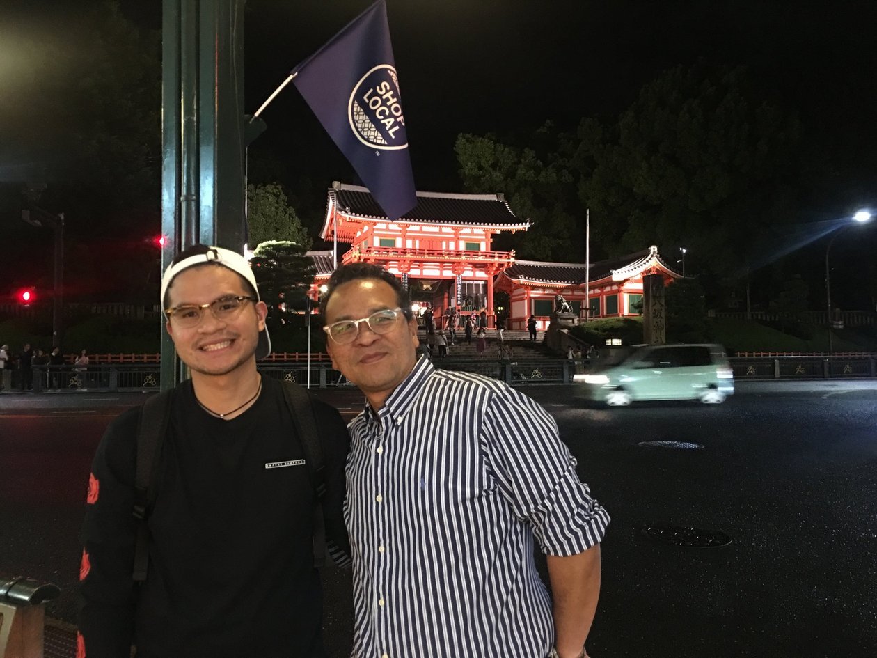 Nico in a black shirt and white cap and Dad in a striped shirt at night, with a lit red shrine gate behind them in Kyoto.