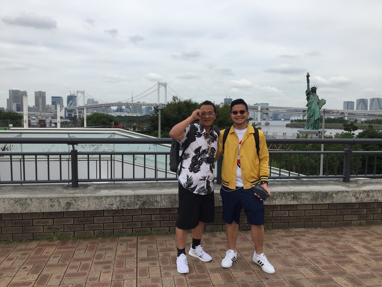 Dad in a leaf-print shirt and Nico in a yellow jacket on a brick overlook, with Rainbow Bridge and the Odaiba Statue of Liberty behind them.