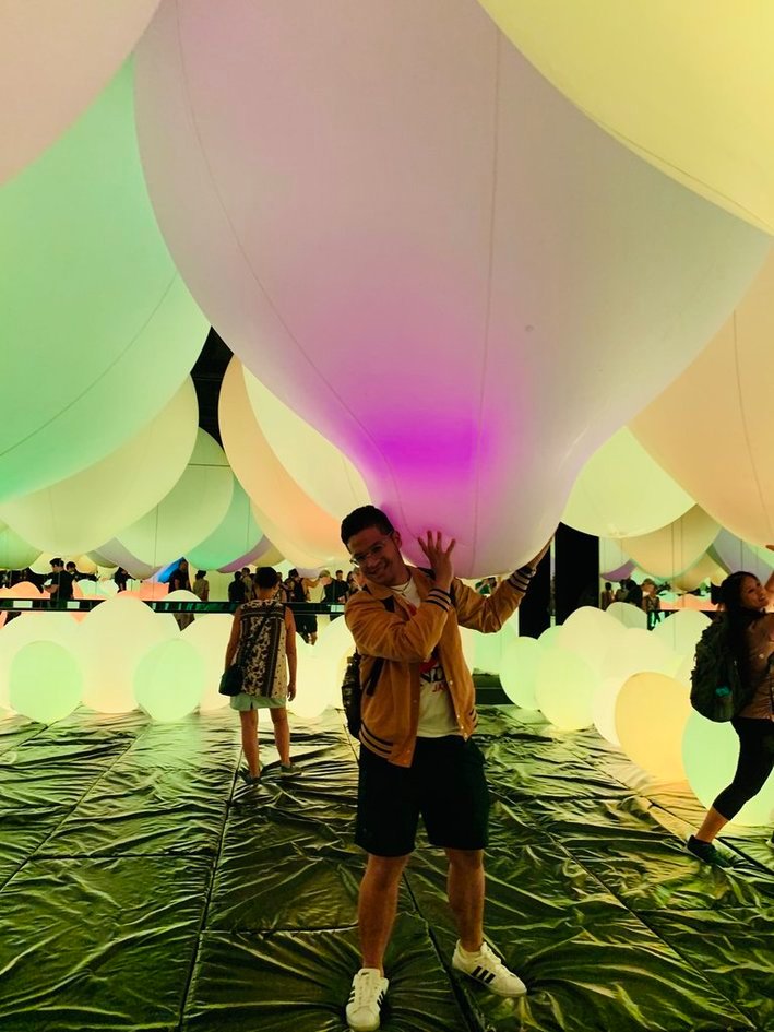 Nico in a yellow jacket holding up a giant glowing balloon in a dark room full of soft colored orbs at teamLab Planets.