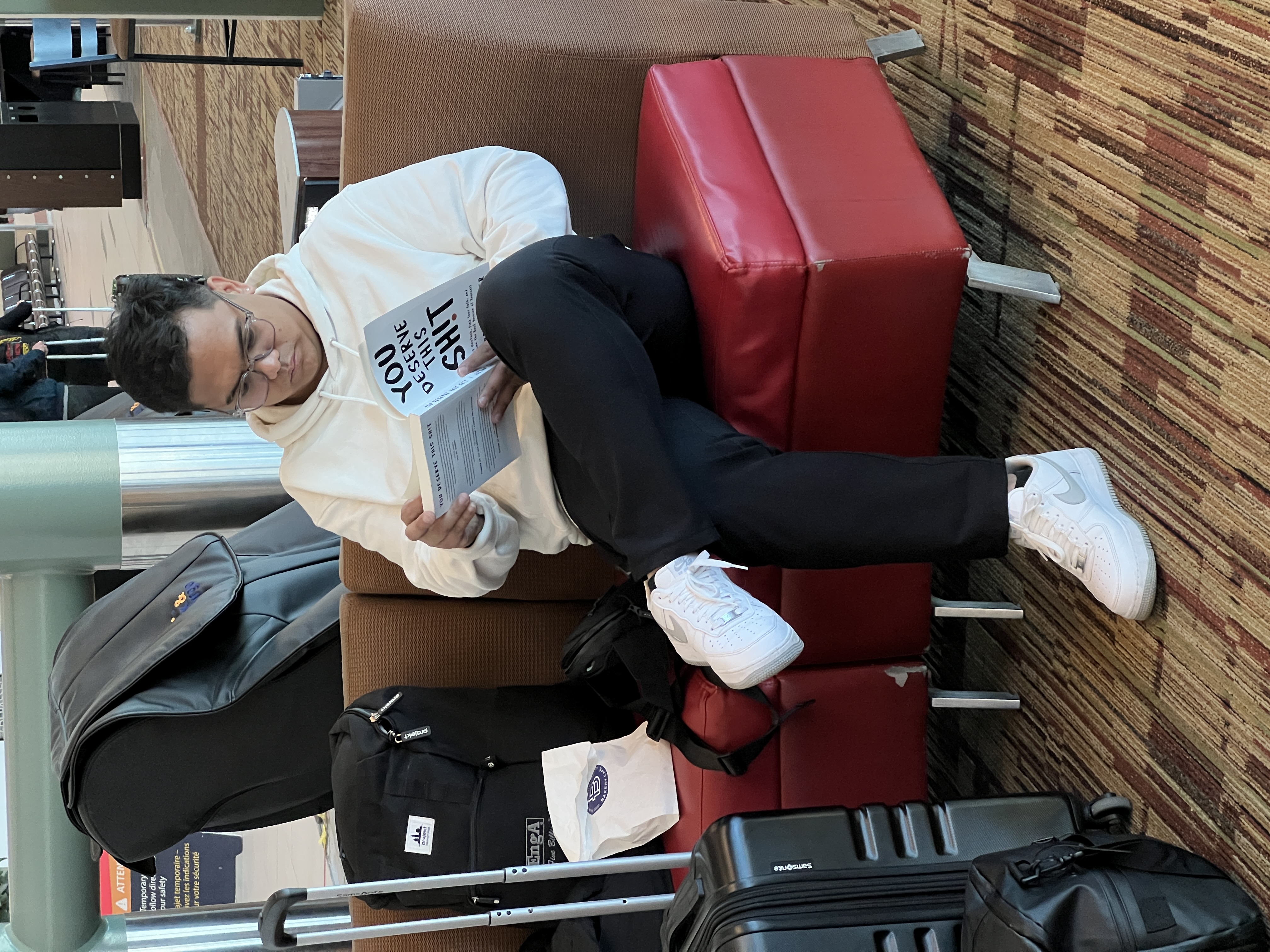 Reading You Deserve This Shit in the airport terminal, white hoodie, glasses, and white sneakers, with luggage on the seats beside him.