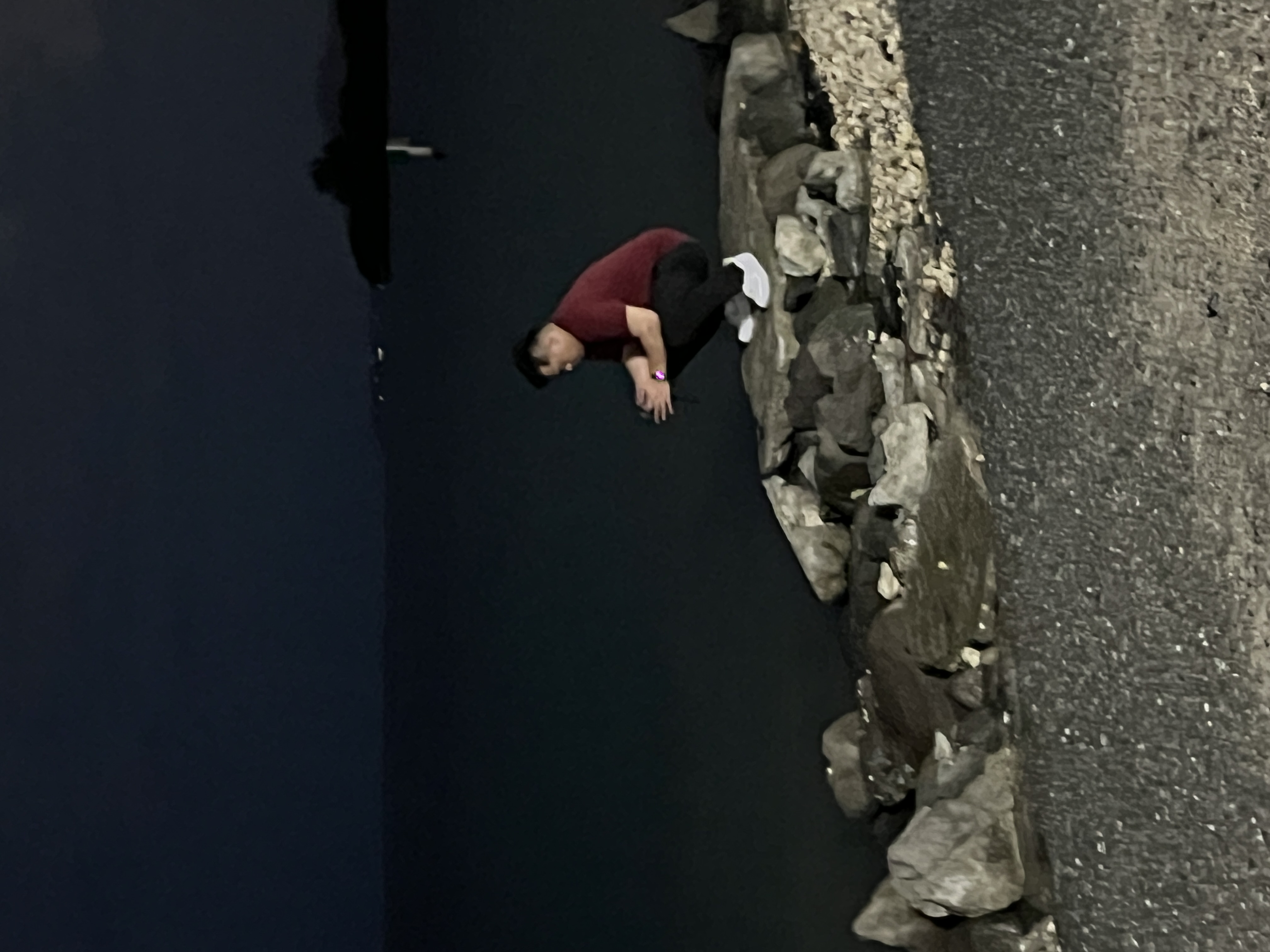 A person in a maroon shirt crouched on the rocks at night at Diamond Head Beach Park, white sneakers, the water behind them.