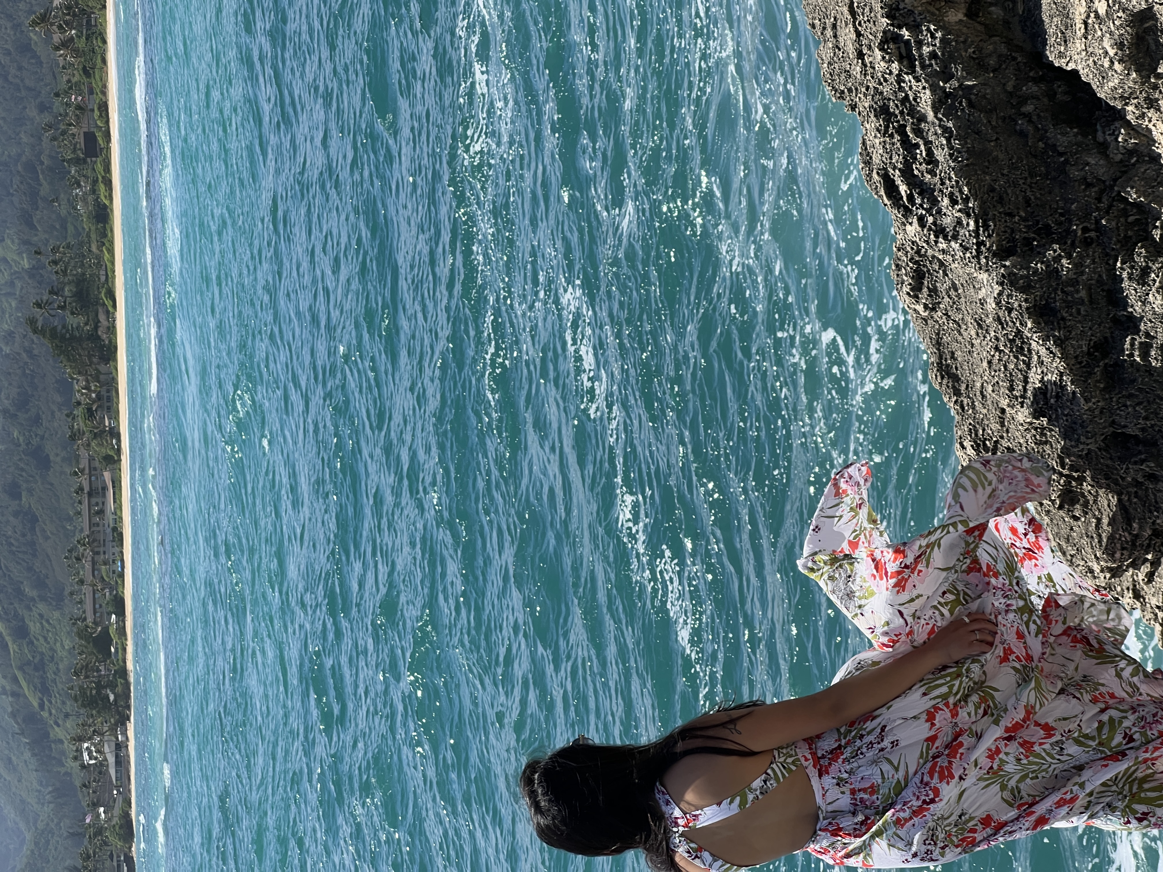 Sarah from behind in the floral dress, standing on the rock and looking out over turquoise water toward a distant beach.