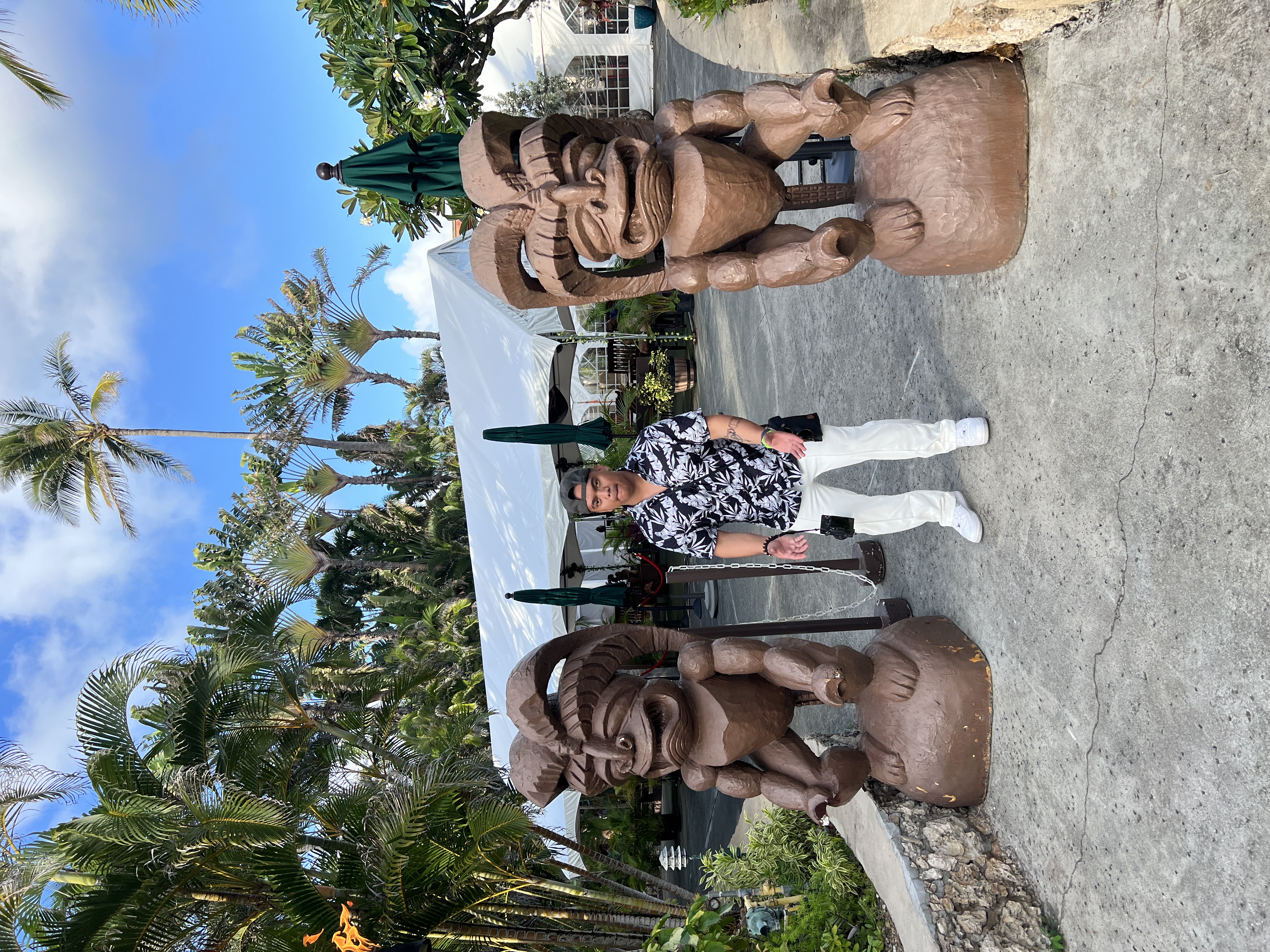 A friend standing between two large carved wooden figures at the Polynesian Cultural Center, white tent and palms behind him.
