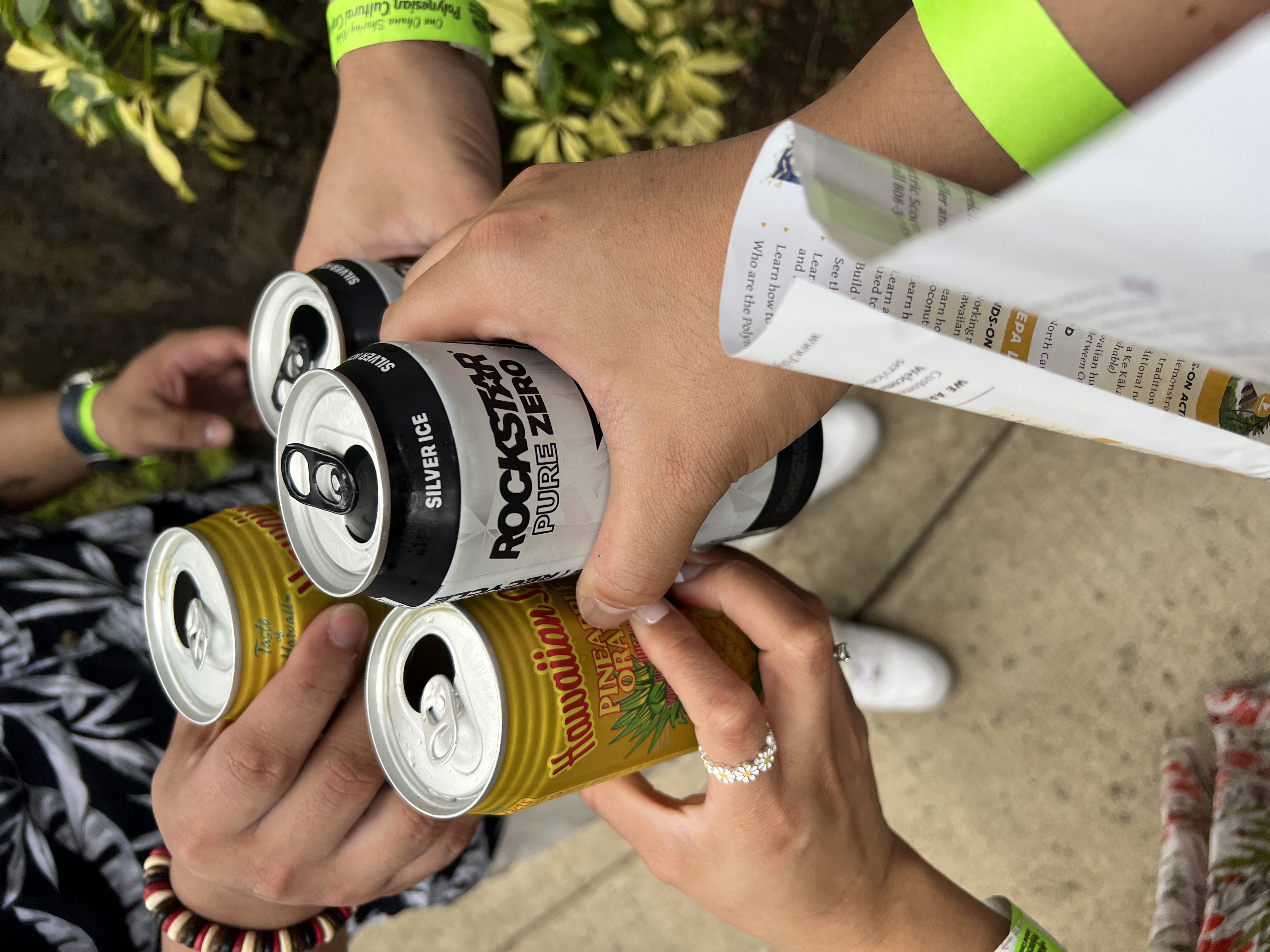 Four hands toasting with Hawaiian Sun pineapple orange cans and a Rockstar, neon Polynesian Cultural Center wristbands visible.