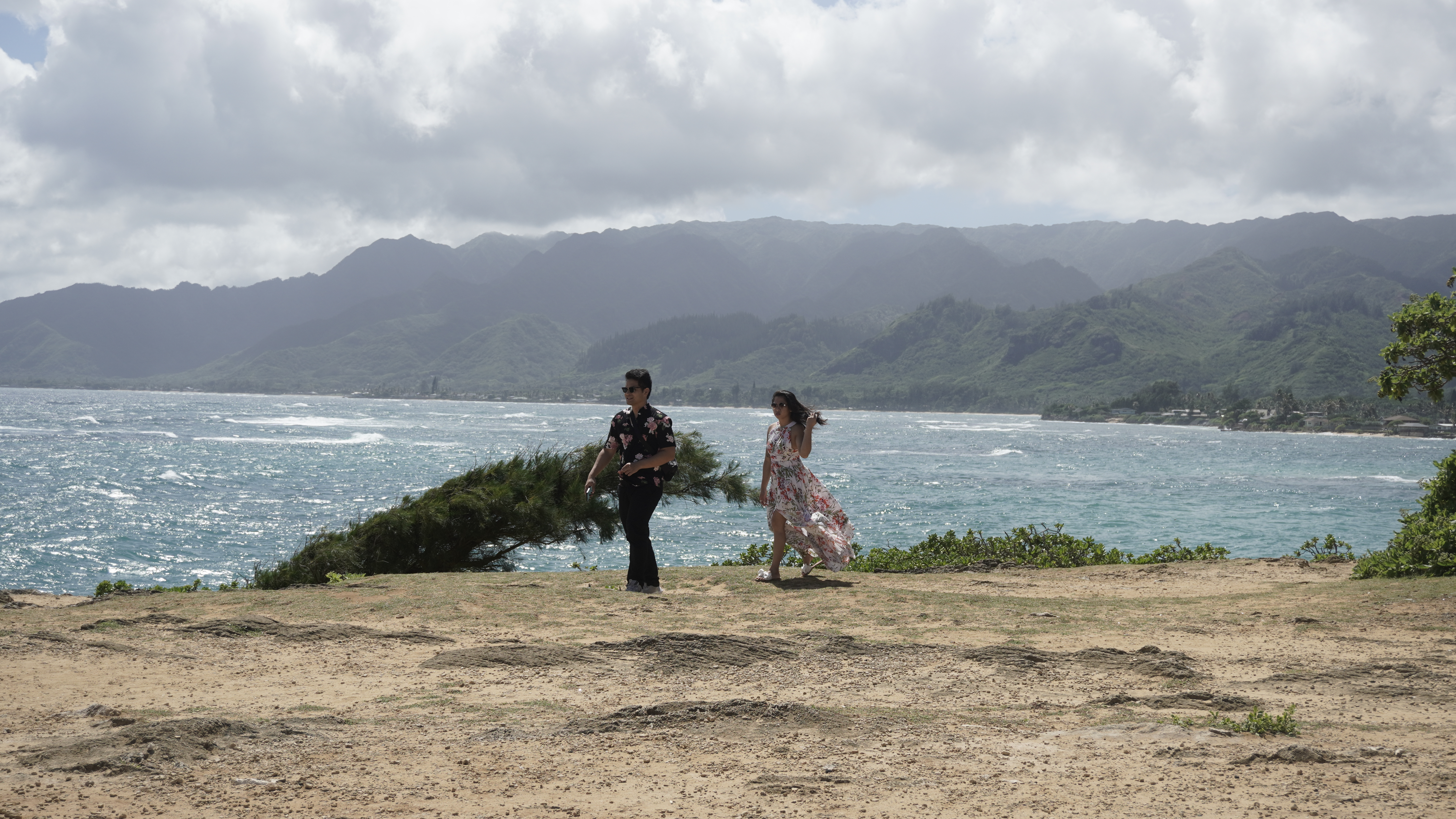 Sarah and Nico walking the dry headland at Lāʻie, the floral dress and black shirt, mountains across the water.