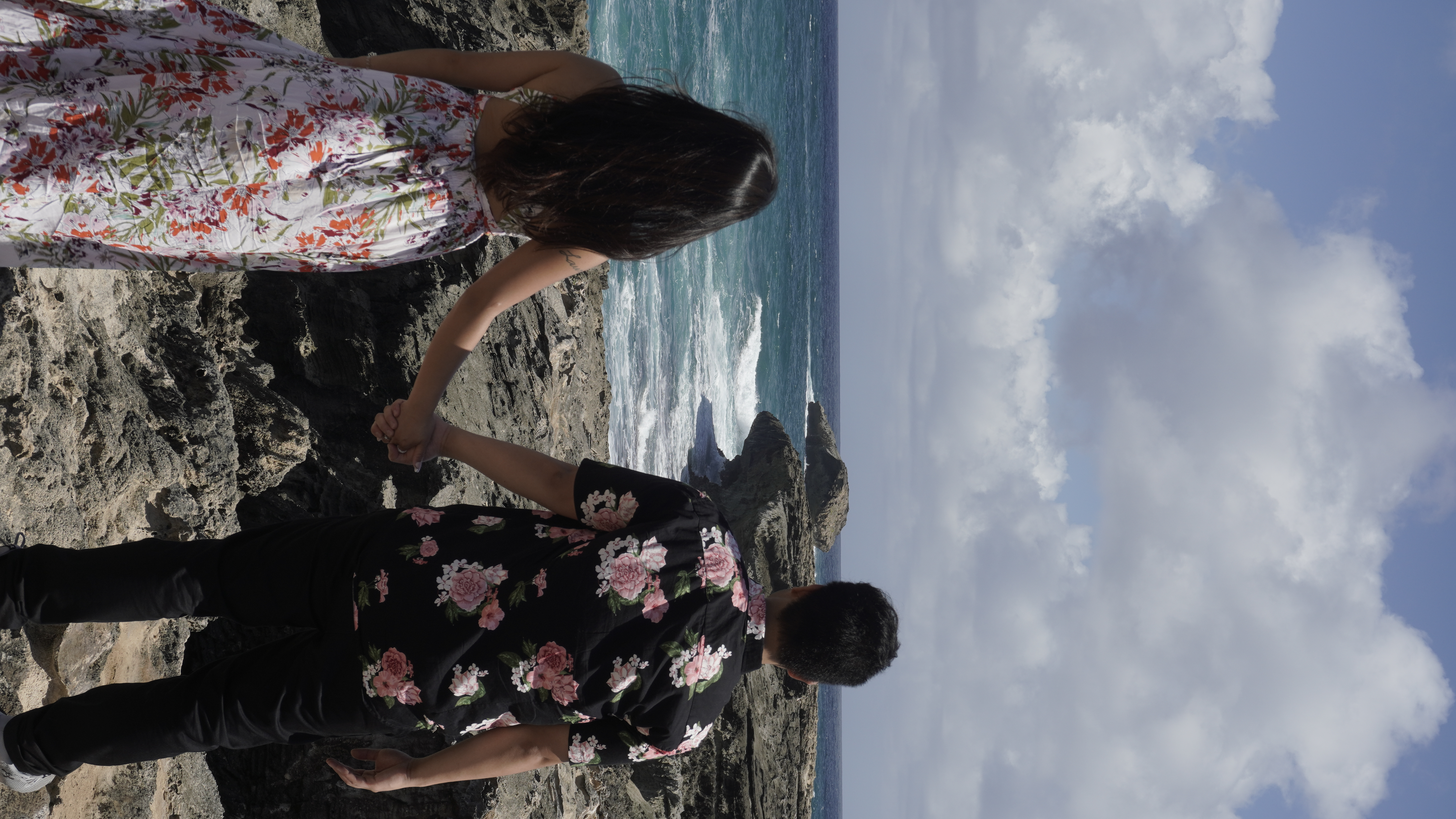 Sarah and Nico from behind, holding hands above a natural rock arch as waves break through it.