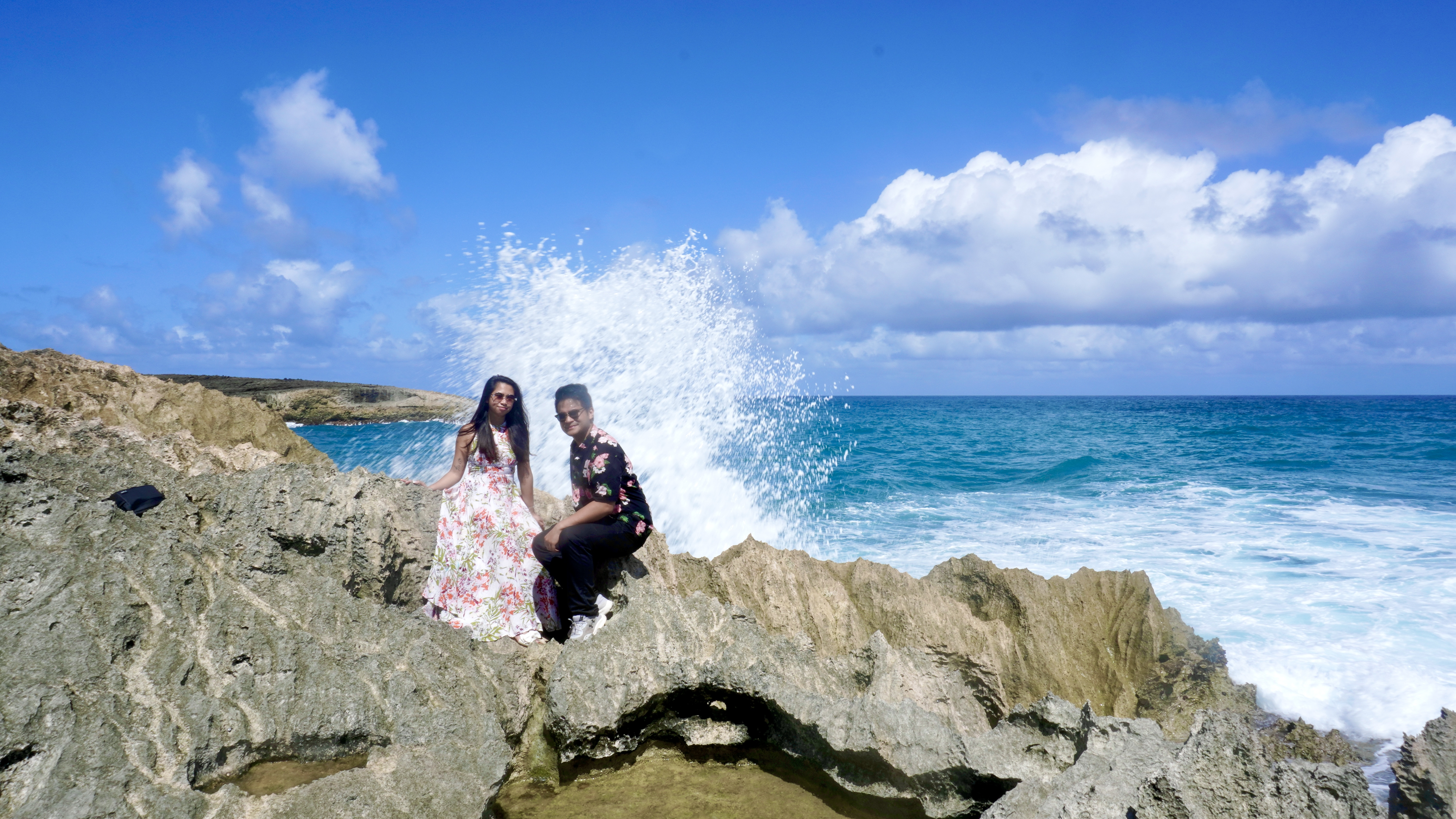 Sarah and Nico sitting on the lava rocks while a wave explodes in white spray behind them.