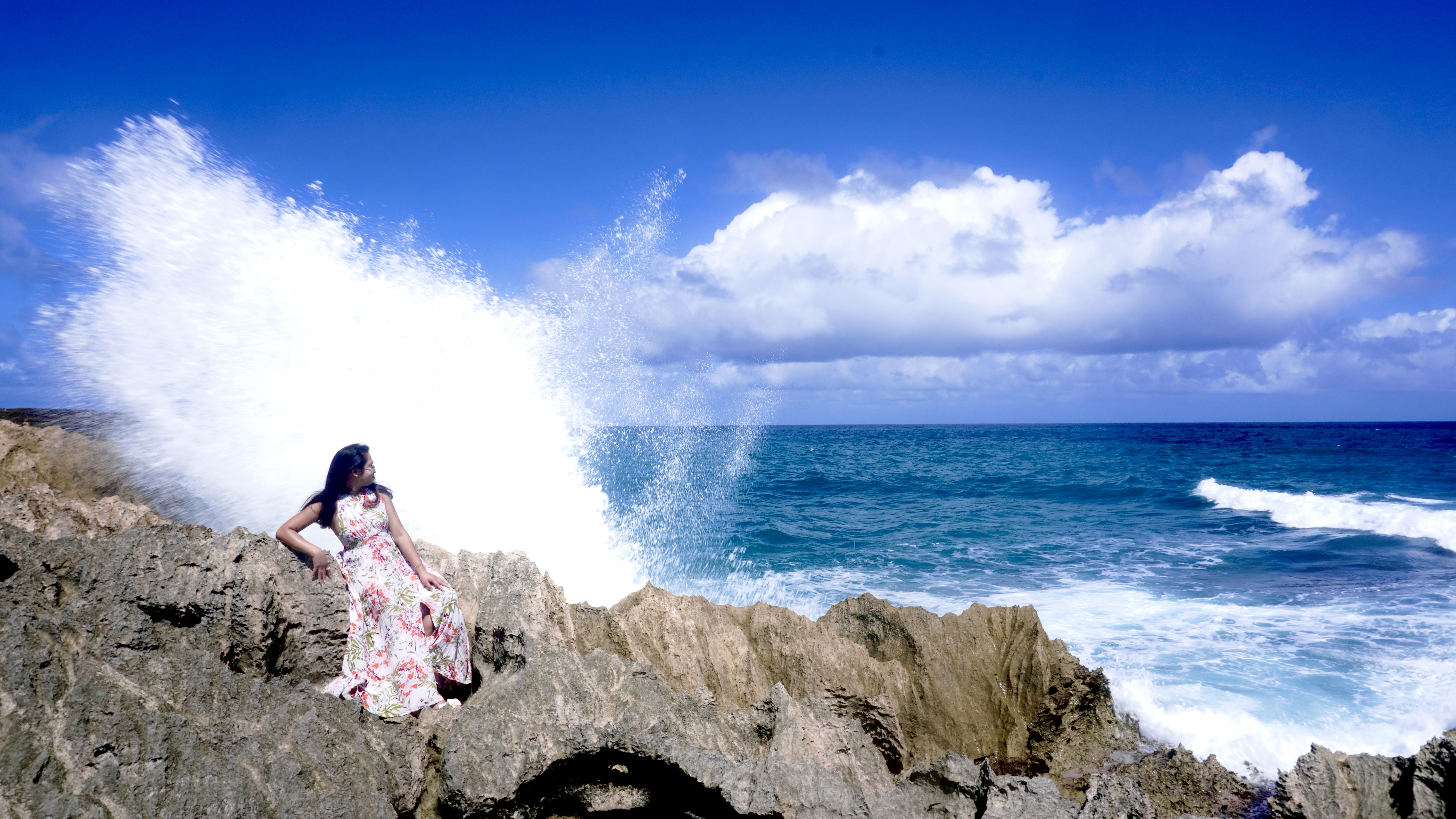 Sarah seated on the rocks in the floral dress as a blowhole throws a tall curtain of spray beside her.