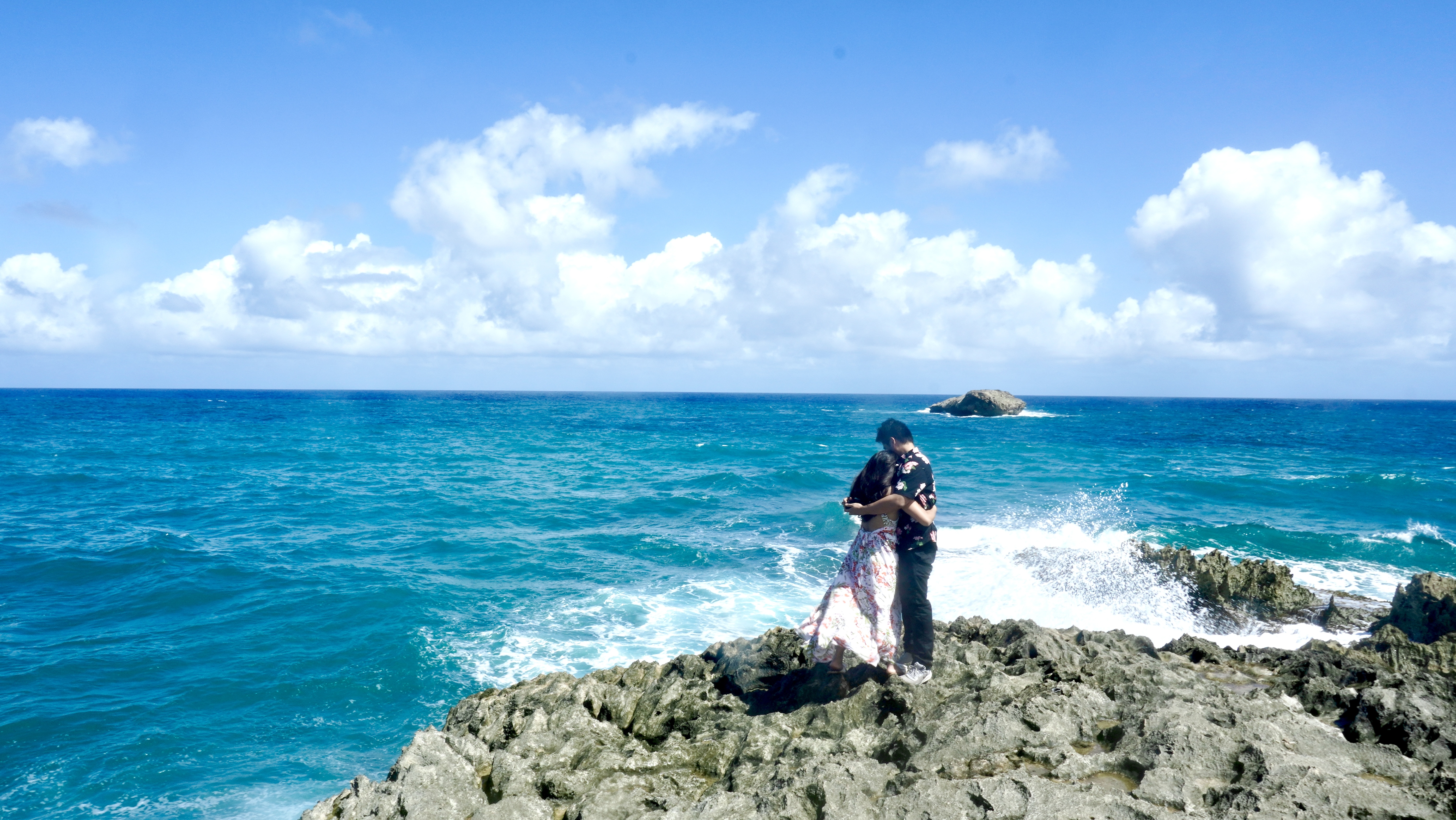 Sarah and Nico embracing on the rocky point, turquoise water and a breaking wave beside them.