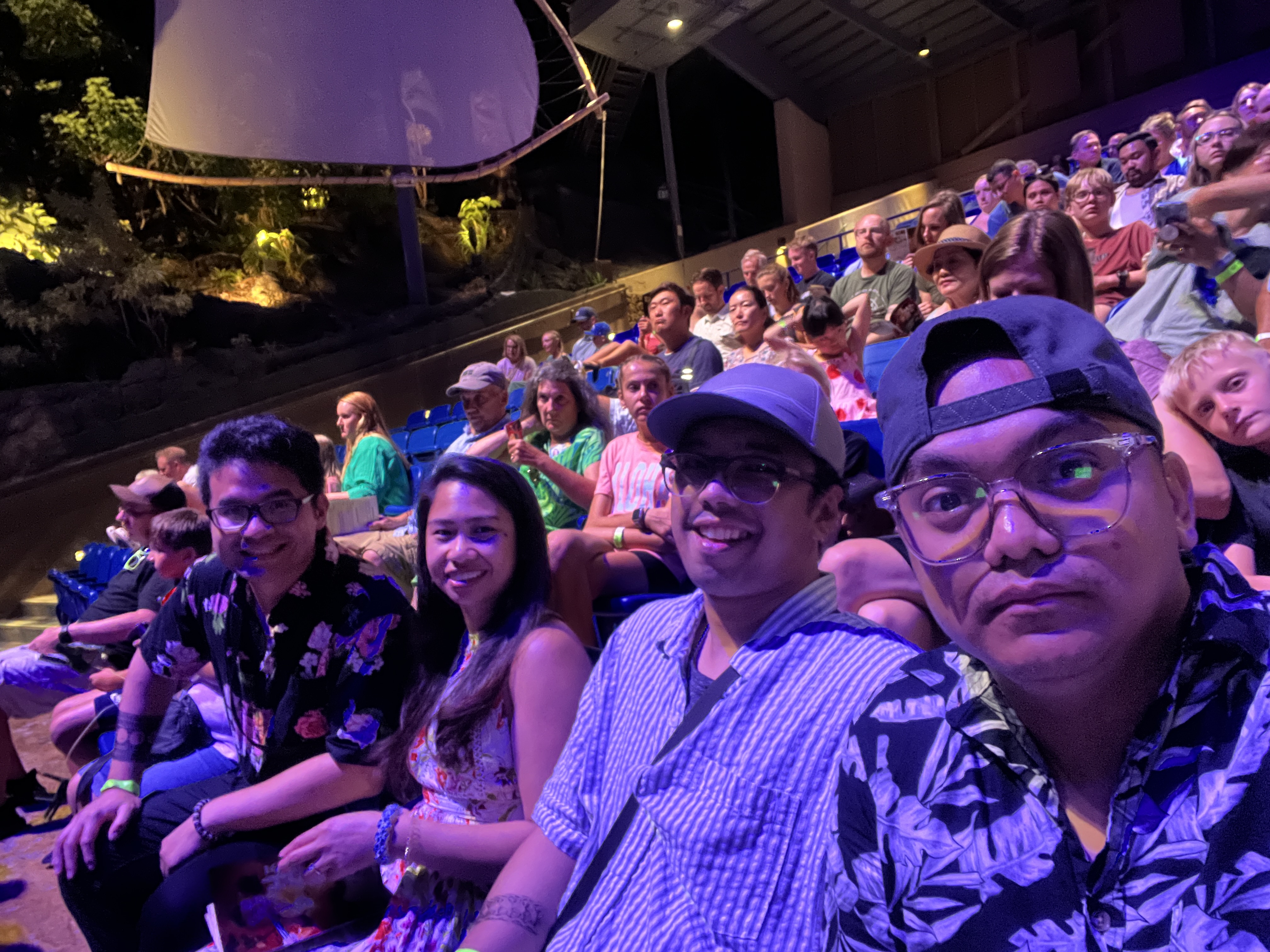 Sarah, Nico, and two friends seated in the audience of the evening show, purple light and a crowded outdoor theater.