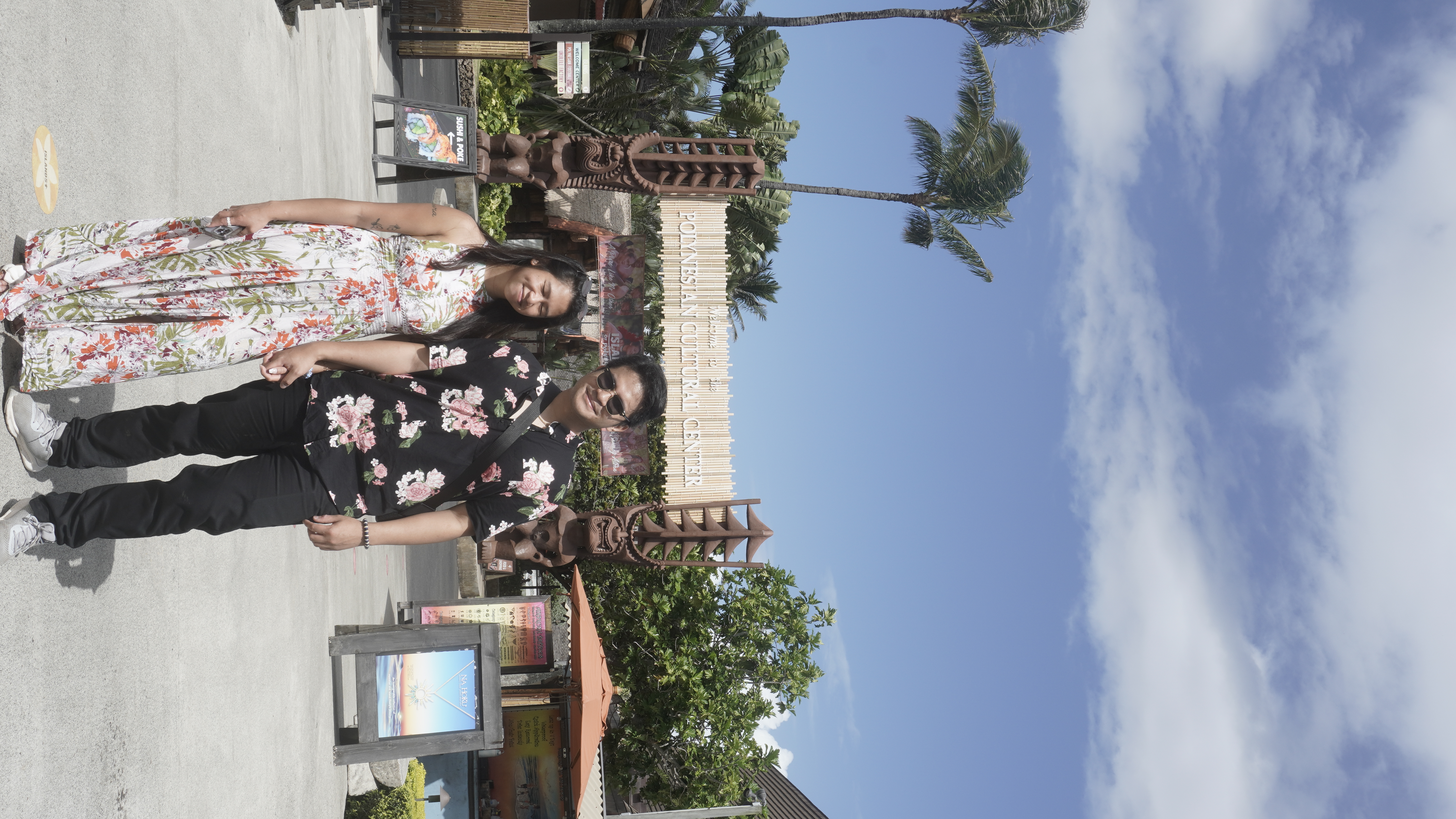 Sarah and Nico holding hands in front of the Polynesian Cultural Center entrance, carved posts and palms around the sign.