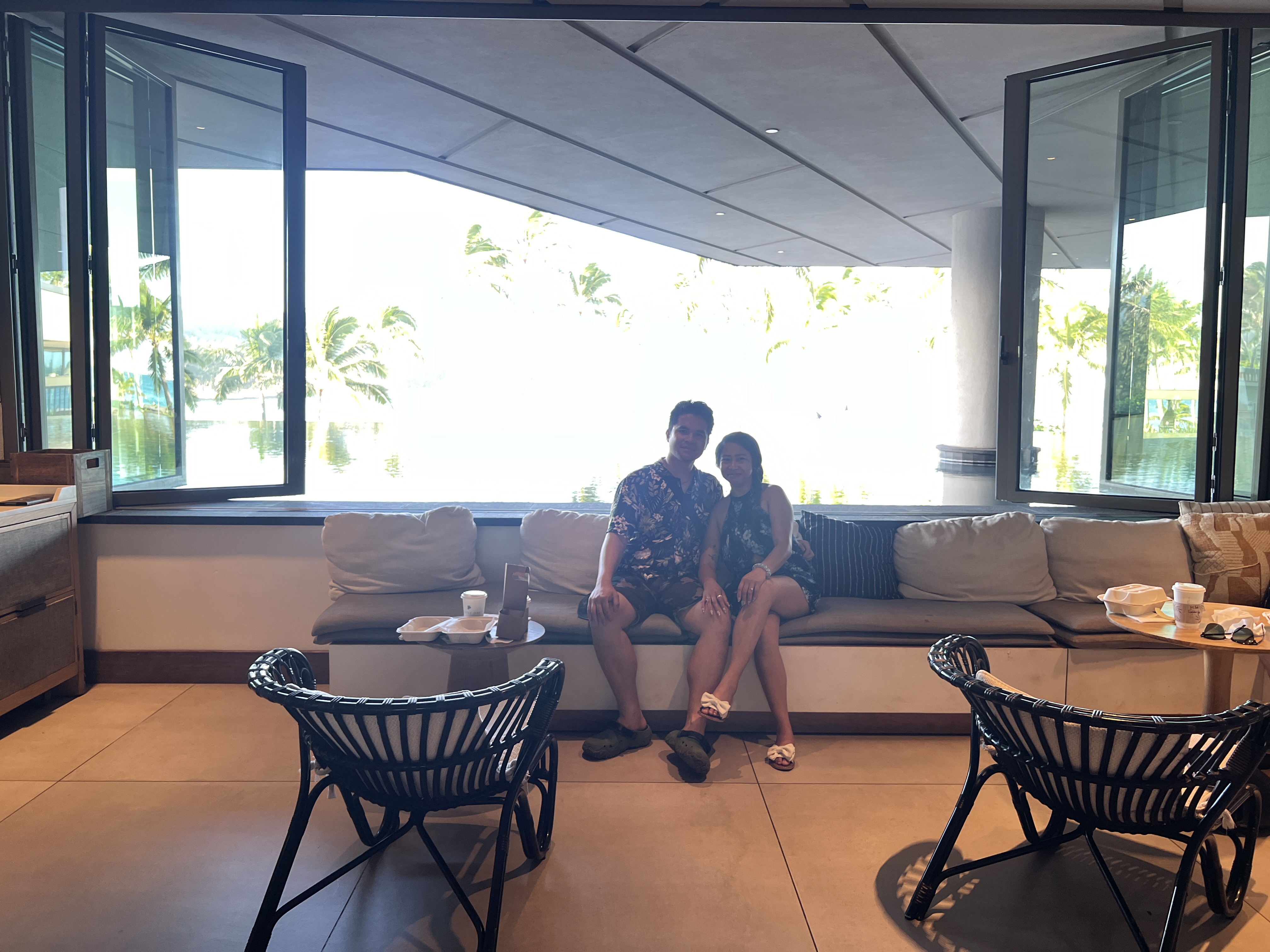 Sarah and Nico on a long bench at a bright resort breakfast, takeout containers beside them and a palm-lined lagoon through the open windows.