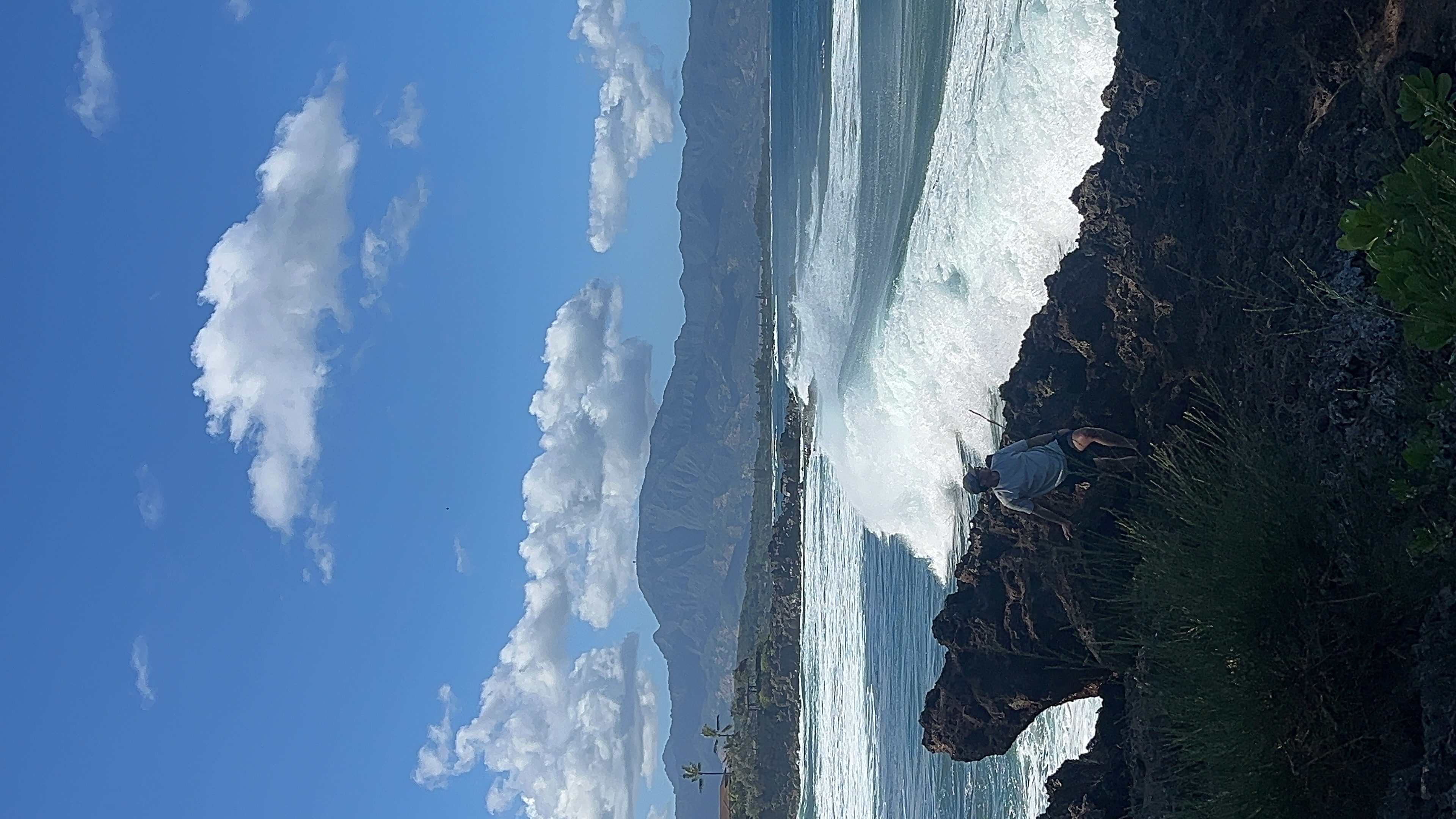 A person sitting on dark lava rock as a large white wave breaks behind them, mountains across the water.