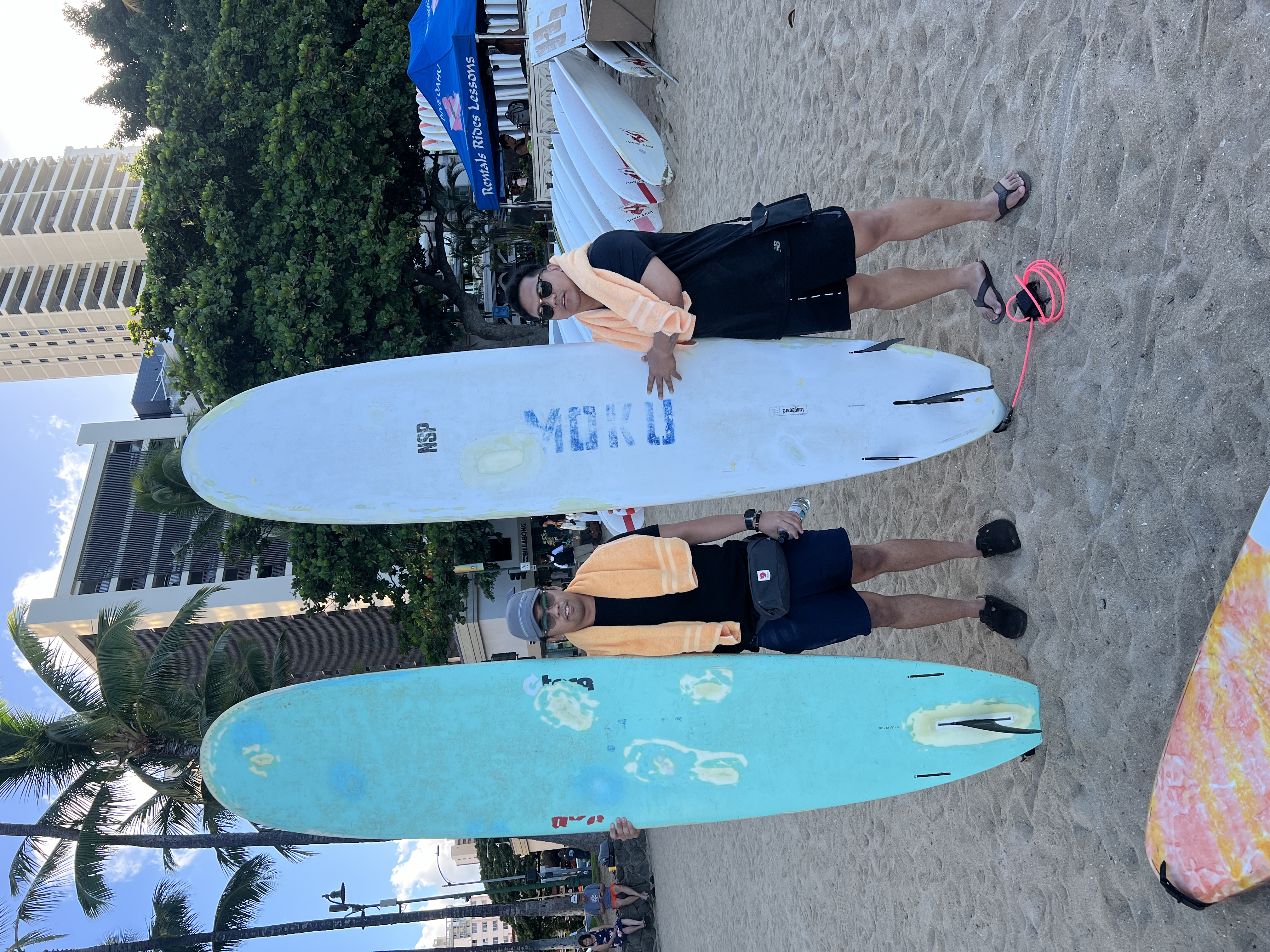 Two friends on Waikiki beach holding long surfboards, orange towels around their necks, a rentals tent and towers behind them.