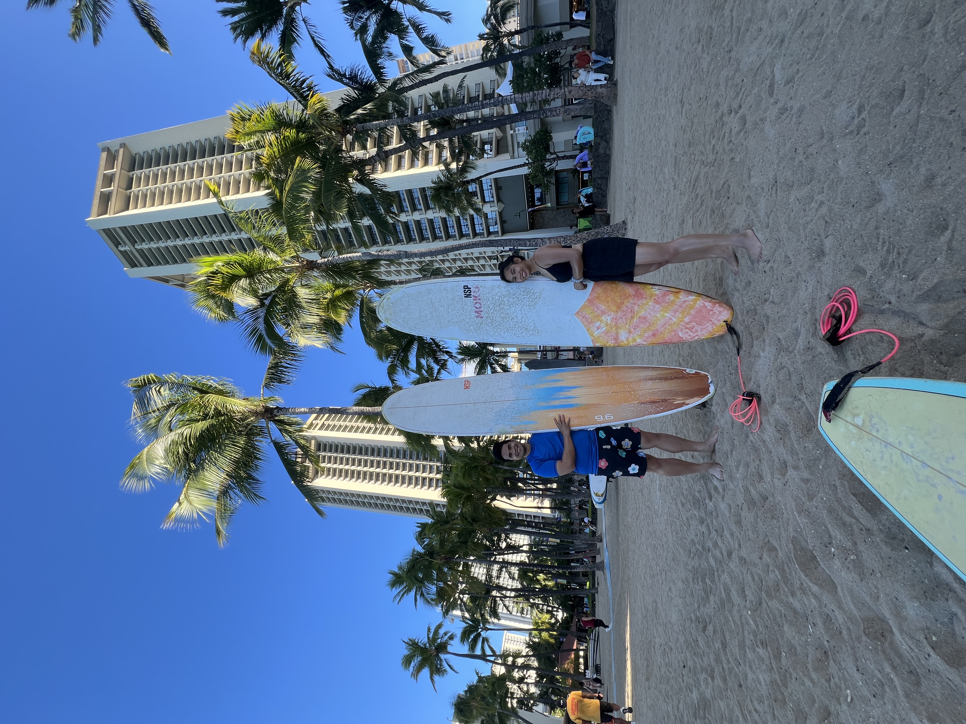 Sarah and Nico standing barefoot with surfboards on Waikiki beach, palms and hotel towers behind them.
