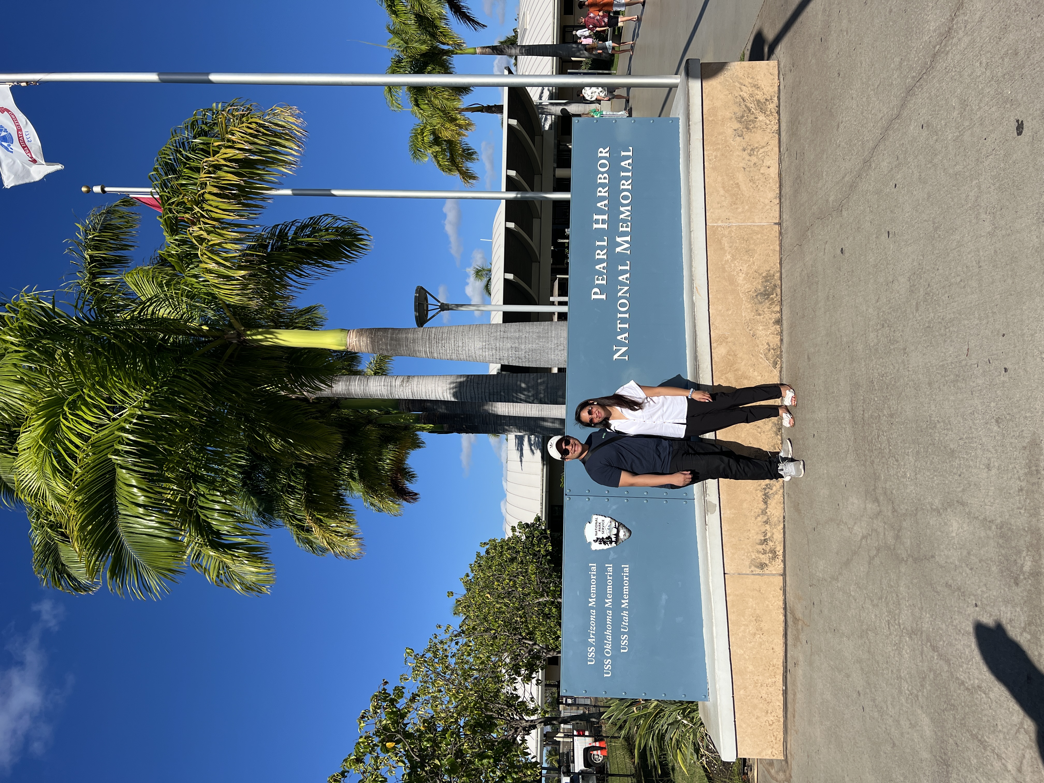 Sarah and Nico standing in front of the blue Pearl Harbor National Memorial sign, palms and flags behind them.