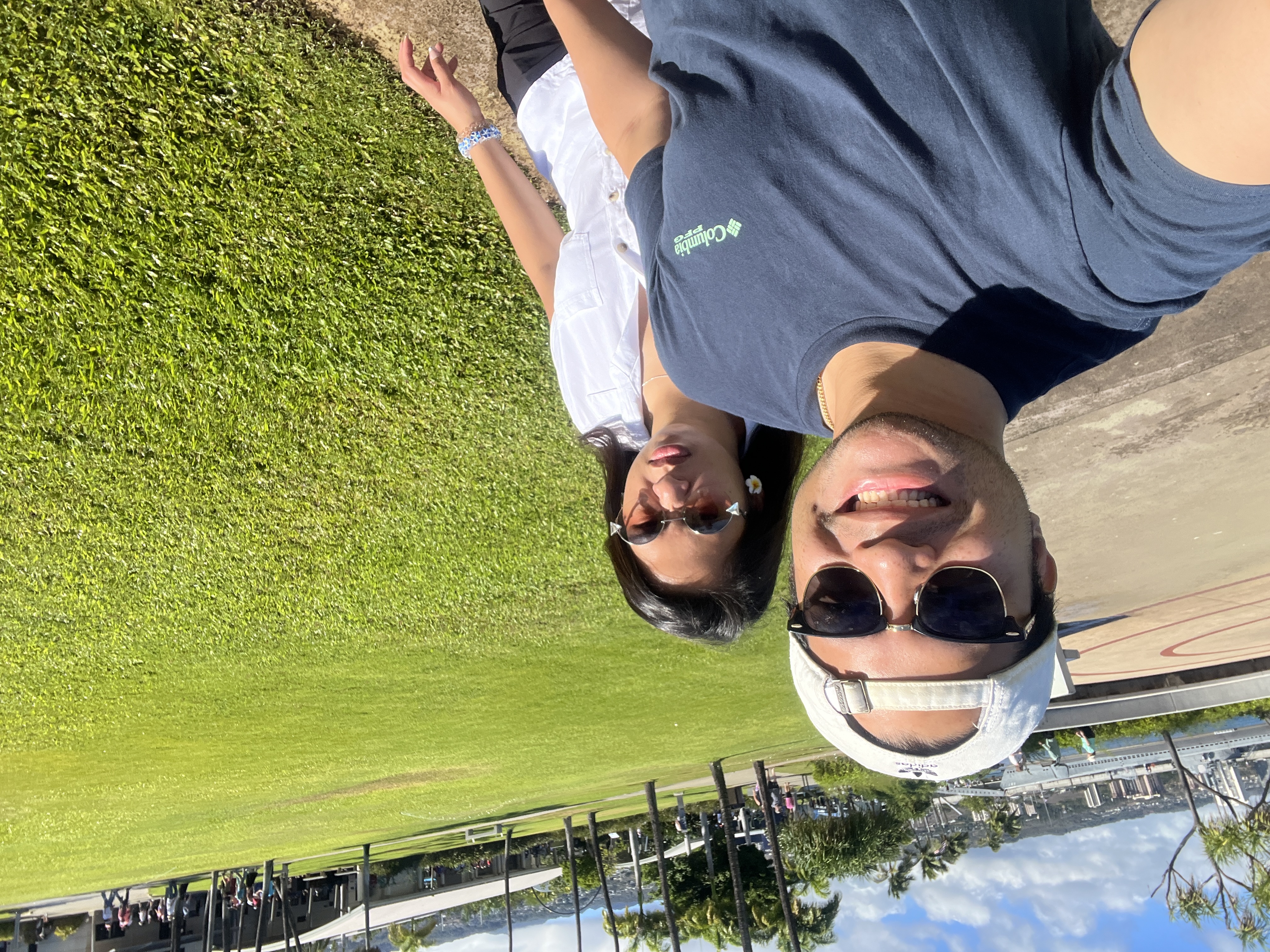 A sunny selfie of Nico and Sarah on the lawn at Pearl Harbor, the harbor and palms behind them.