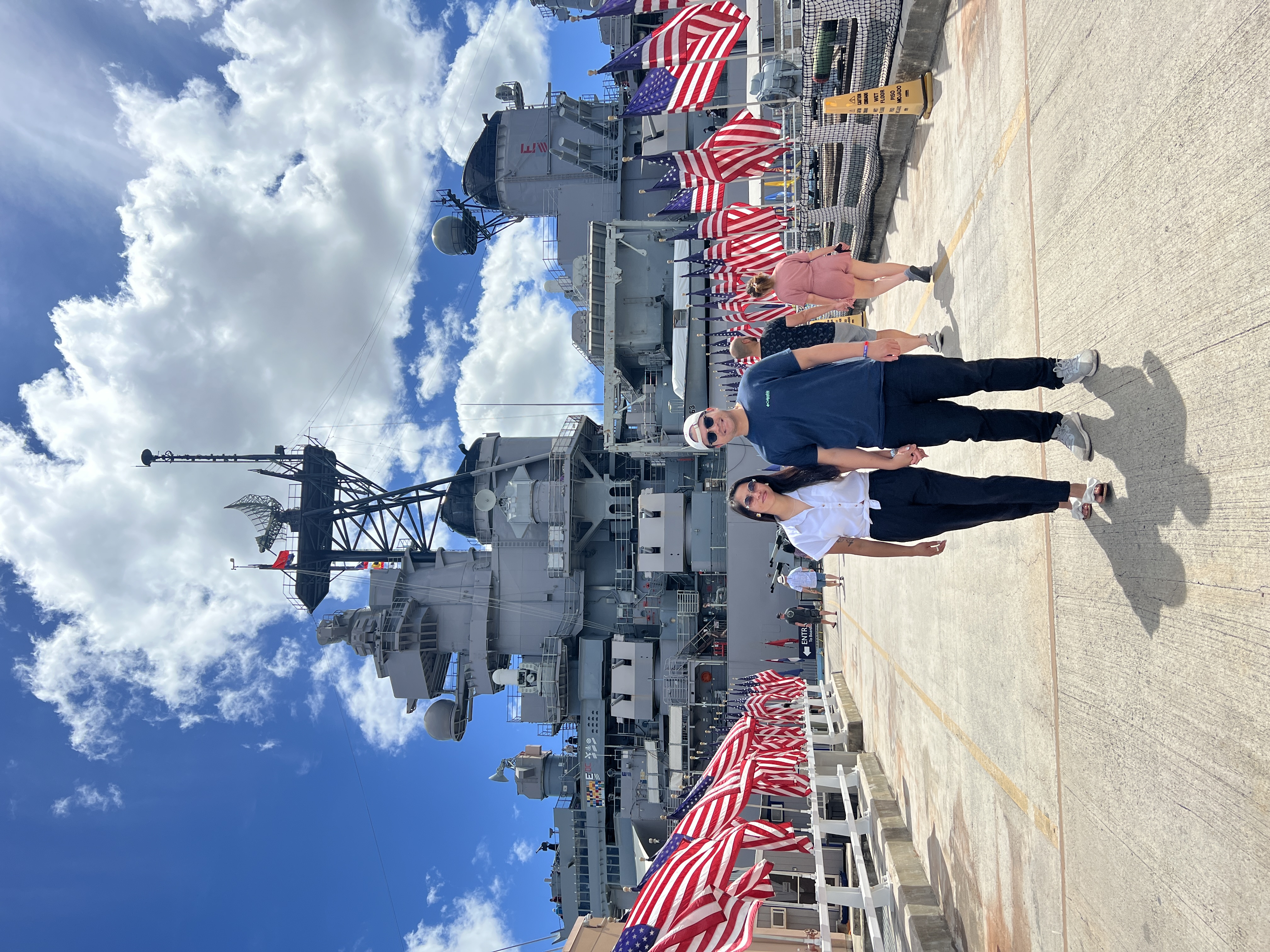 Sarah and Nico holding hands on the deck of the USS Missouri, rows of American flags leading toward the ship.