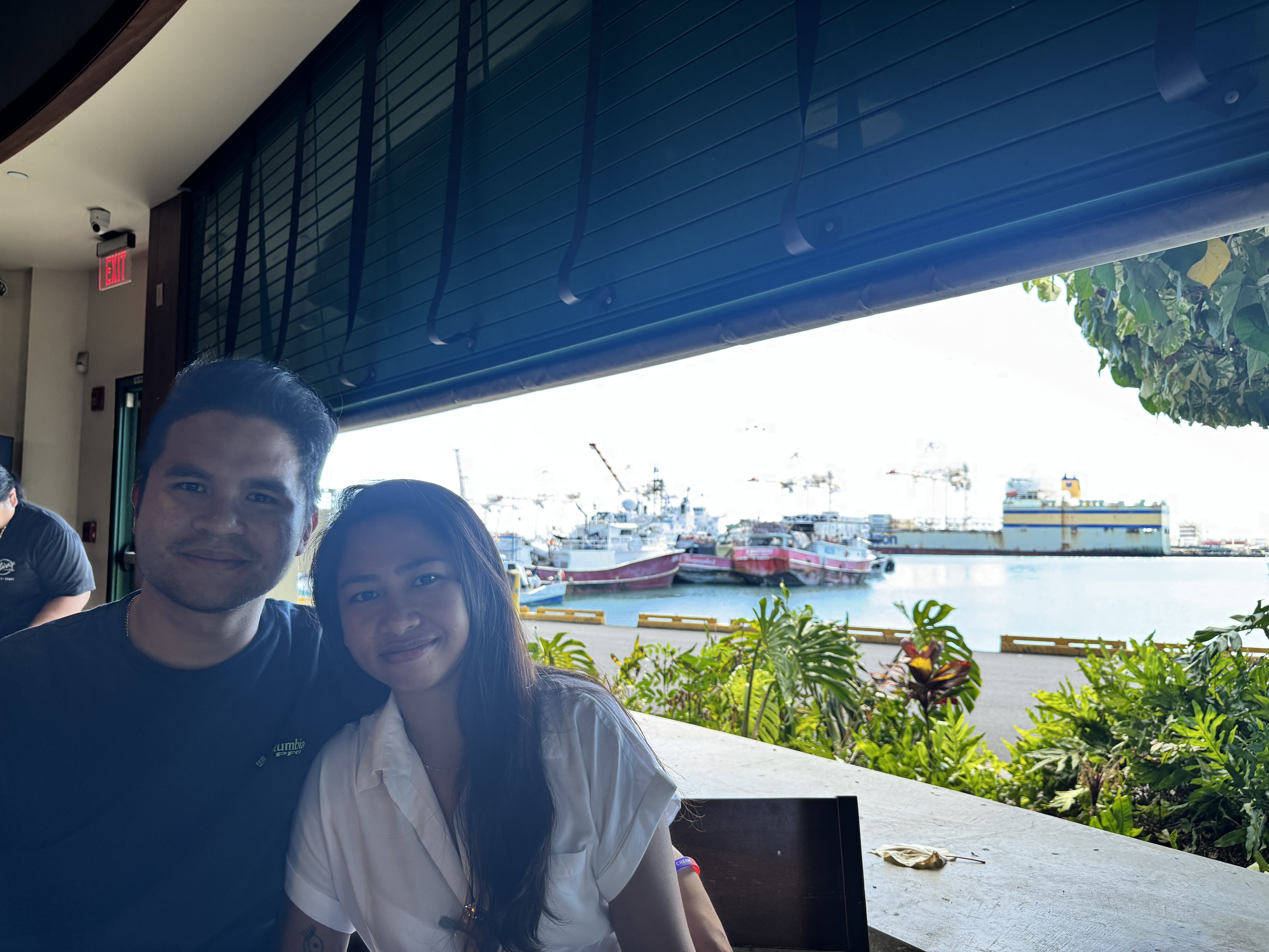 Sarah and Nico at a table beside an open waterfront awning, fishing boats and a large ship in the harbor behind them.