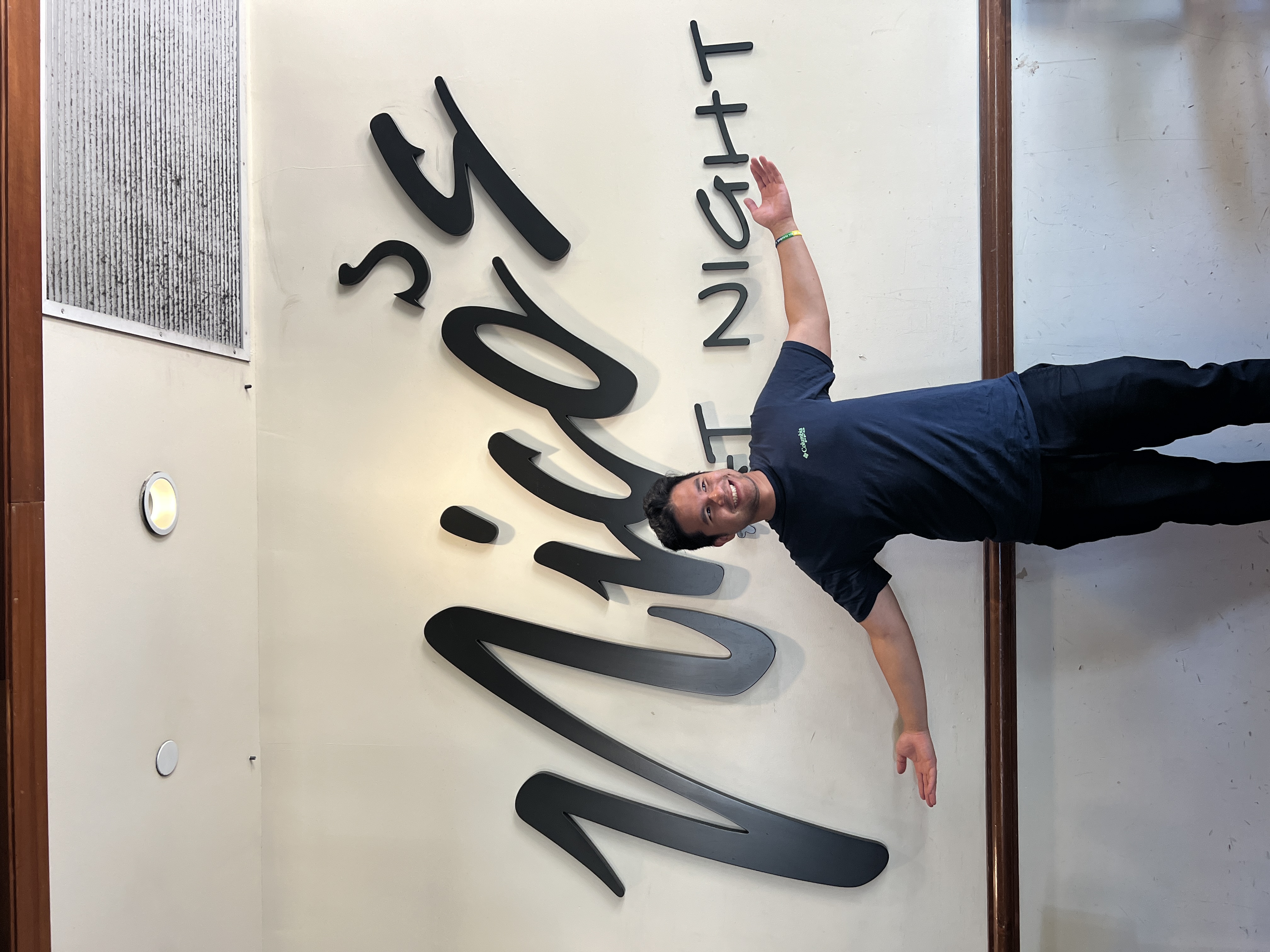 Nico standing with arms out under a large black Nico’s sign on a restaurant wall.