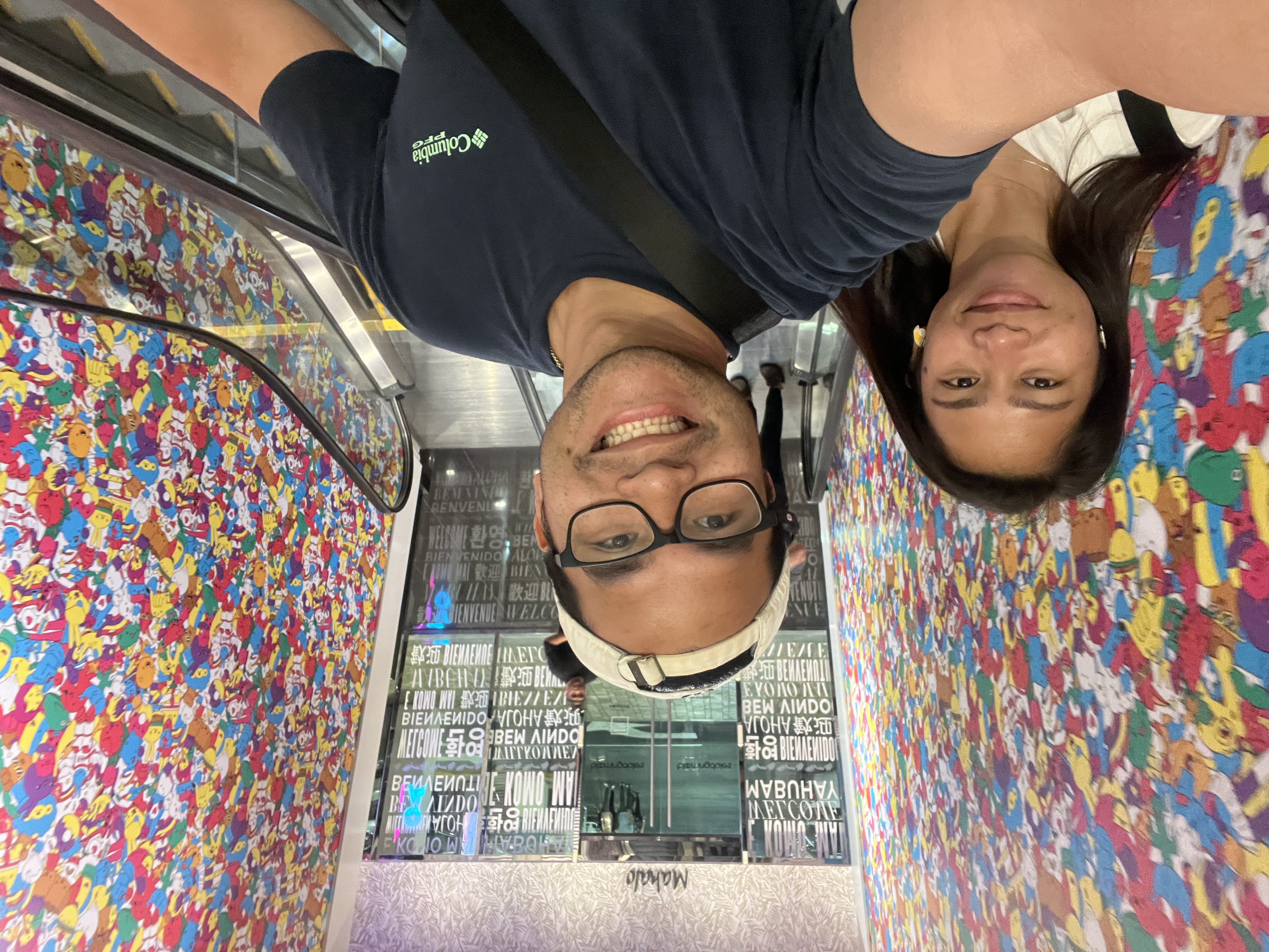 Sarah and Nico on an escalator beneath a Mahalo sign, colorful wallpaper and welcome written in many languages.